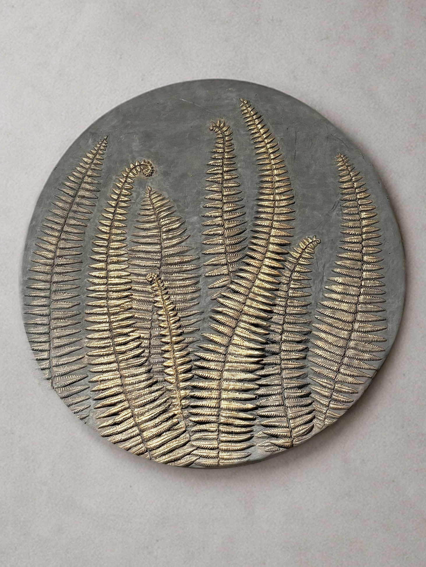A round gray decorative wall plaque featuring embossed metallic gold fern leaves, shown against a neutral light gray background daba deco